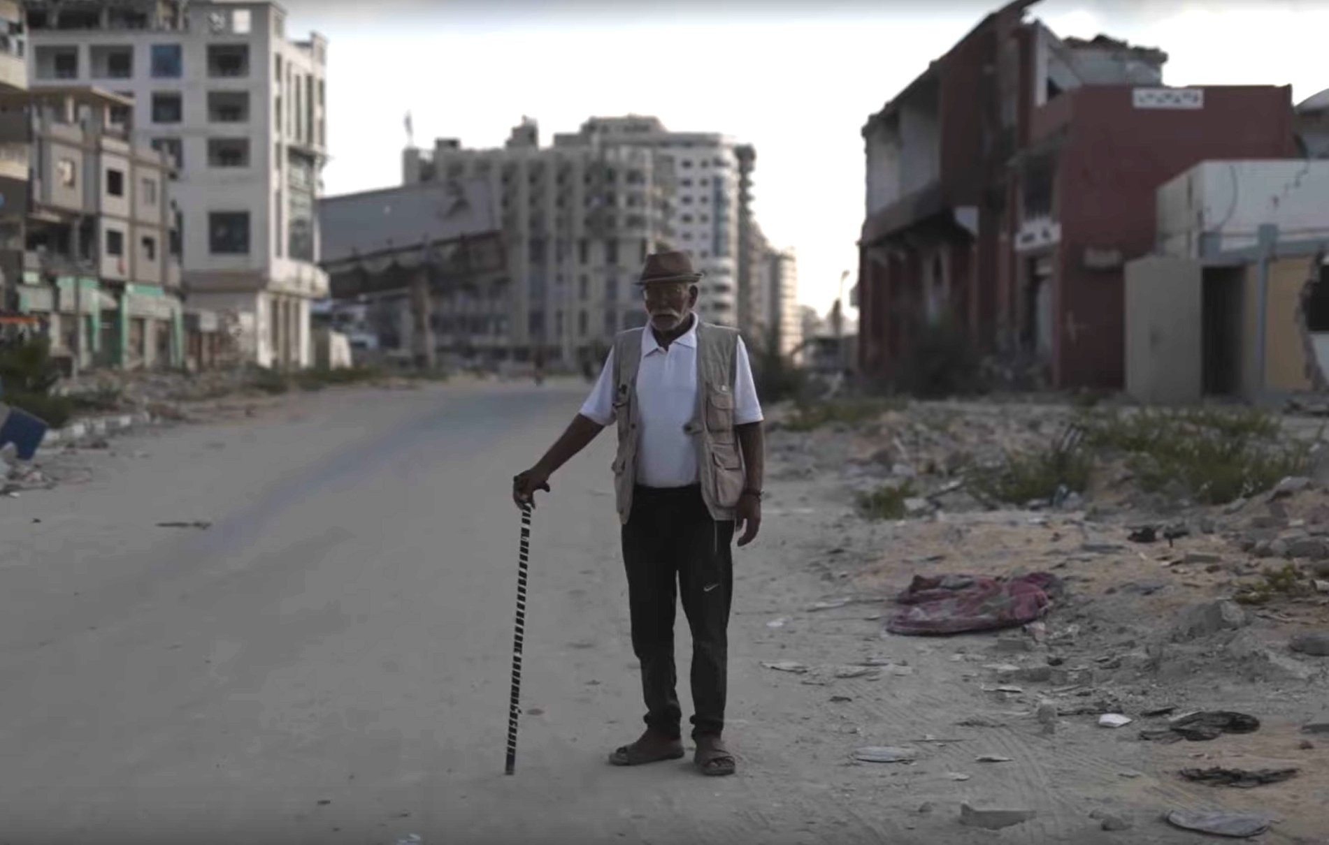 Un homme âgé avec une canne se tient sur une rue déserte, entouré de bâtiments en ruine.