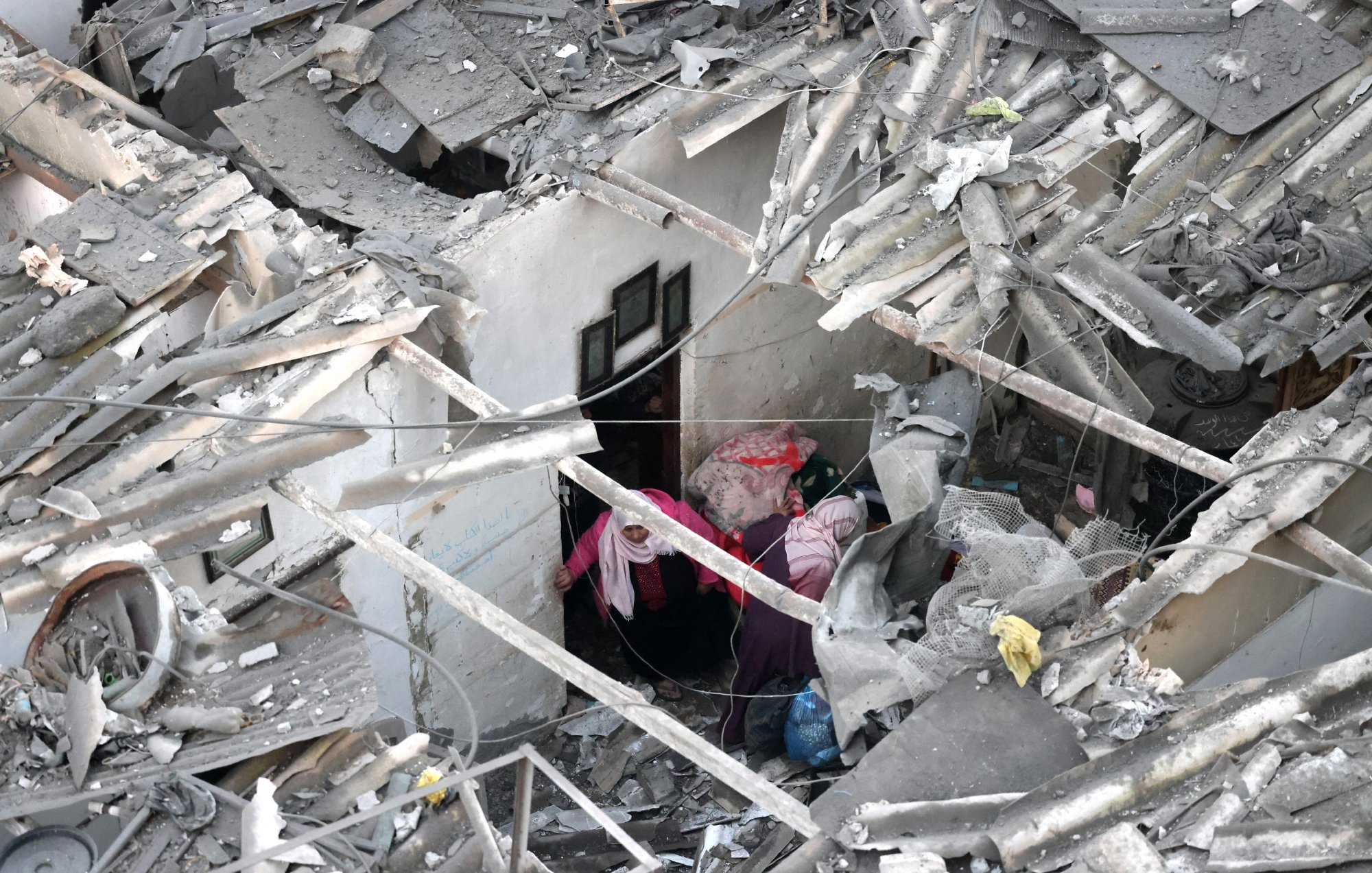 L'image montre des ruines d'un bâtiment, avec des débris de toit et des murs effondrés. Deux personnes sont visibles à l'intérieur des décombres, semblant chercher ou s'entraider dans cet environnement chaotique.