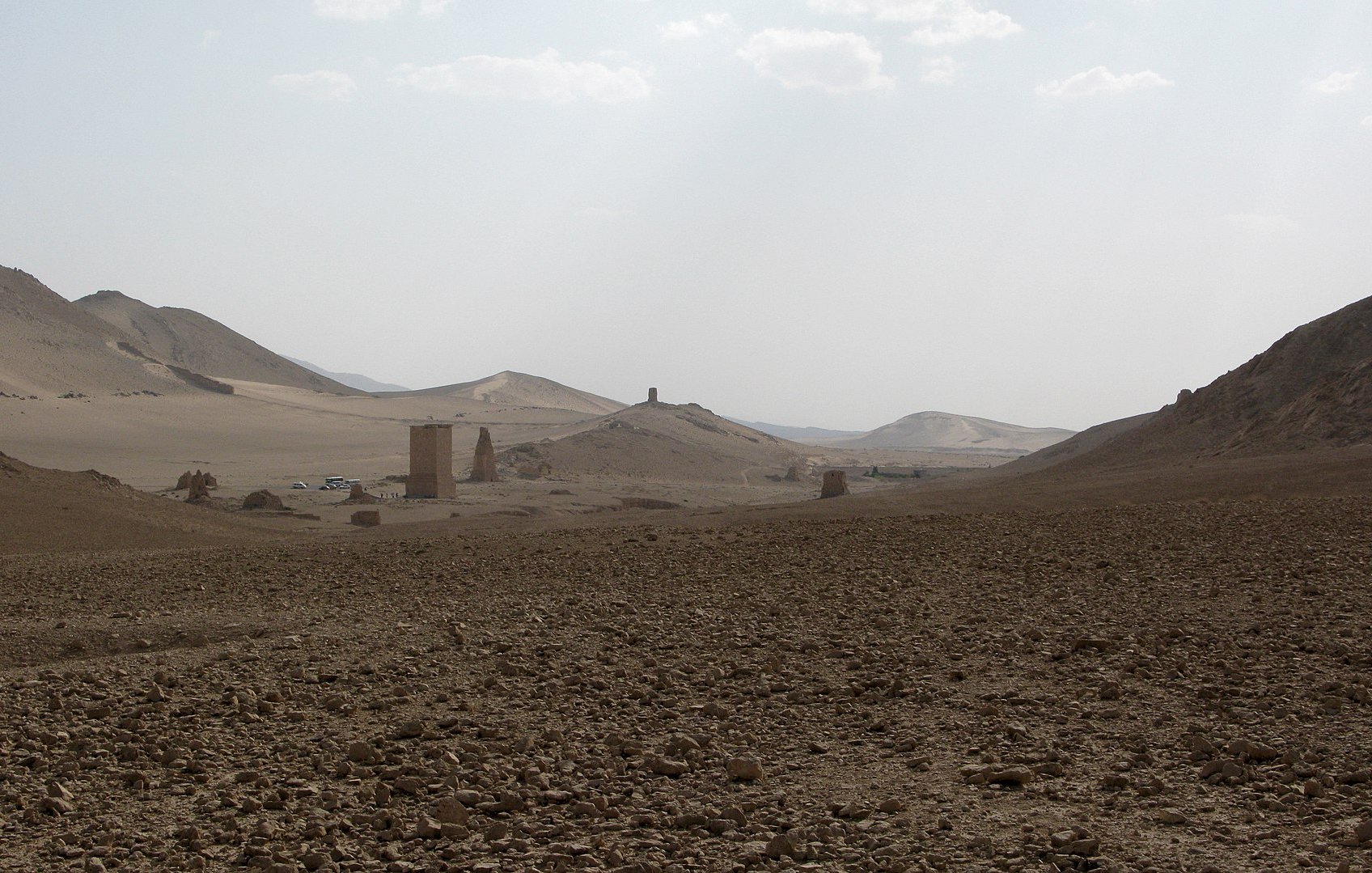 L'image montre un paysage désertique avec des collines et des formations rocheuses. Le sol est aride, parsemé de cailloux et de petites pierres. À l'horizon, on peut apercevoir quelques structures anciennes, probablement des vestiges ou des ruines, qui se détachent dans le paysage. Le ciel est clair, avec quelques nuages, et la lumière semble douce, créant une ambiance tranquille et isolée.