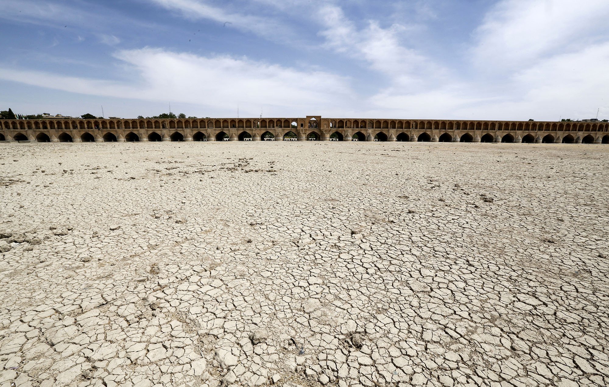 L'image montre un paysage aride avec des sols craquelés, indiquant une sécheresse sévère. À l'arrière-plan, on aperçoit un grand pont historique avec plusieurs arches. Le ciel est dégagé avec quelques nuages, et l'absence d'eau dans la zone suggère des conditions environnementales préoccupantes. Cette scène illustre les effets de la déshydratation sur un espace habituellement aquatique.