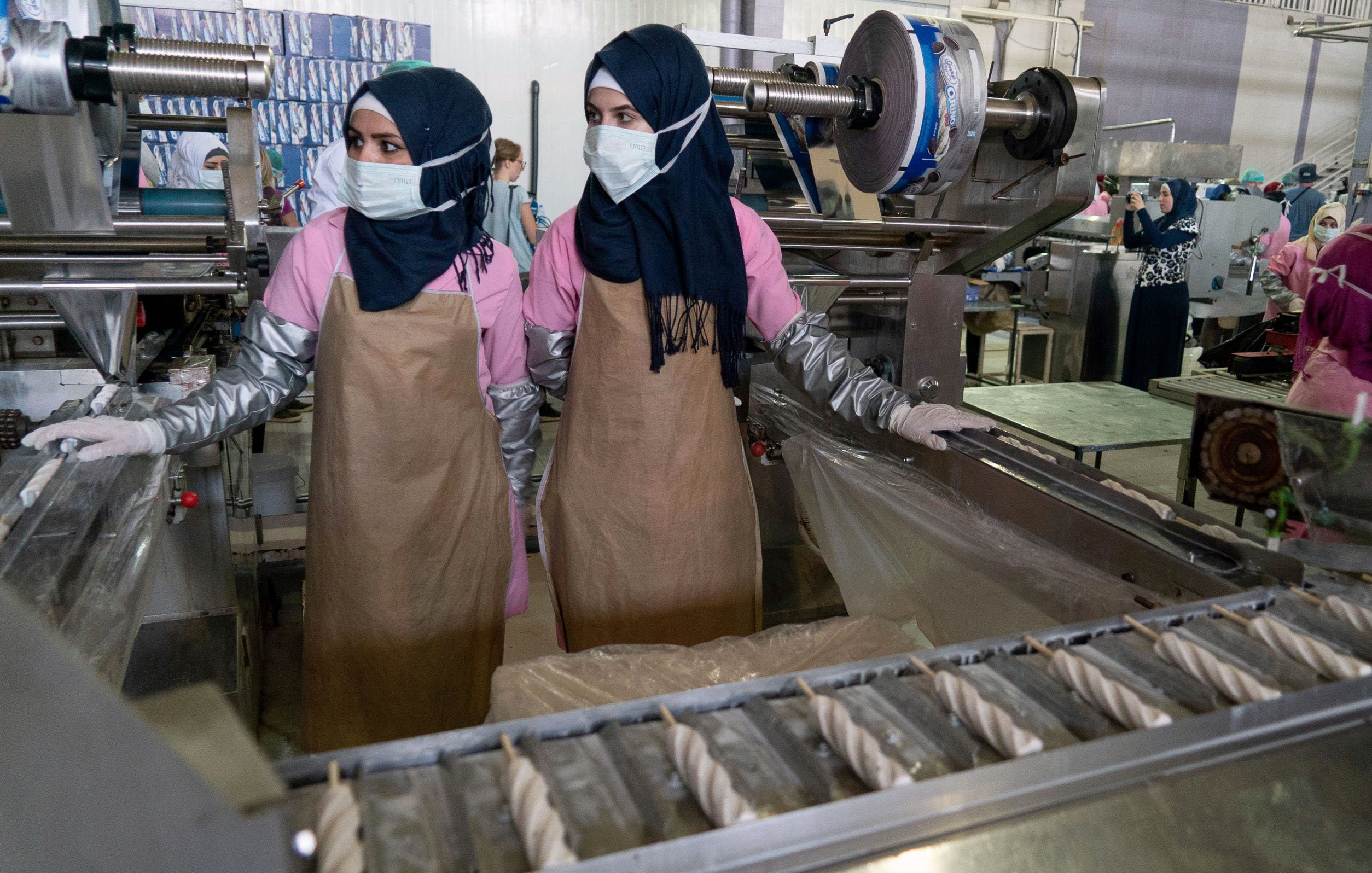 L'image montre deux femmes travaillant dans un environnement industriel. Elles portent des hijabs et des masques de protection. Elles sont habillées de vêtements de travail, avec des blouses et des tabliers. À l'arrière-plan, on peut voir des machines et d'autres personnes également en train de travailler, ce qui indique qu'il s'agit probablement d'une usine ou d'un atelier de production. L'atmosphère semble axée sur le travail et la production.