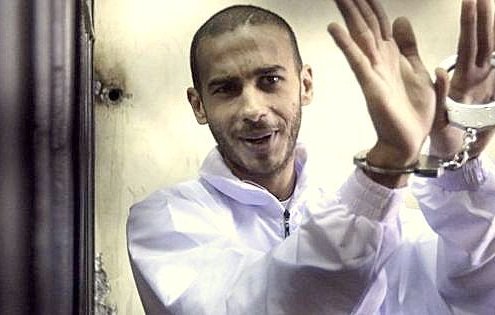 L'image montre un homme au regard confiant, portant une combinaison blanche. Ses mains sont menottées, et il semble faire un geste de la main, peut-être en souriant ou en se moquant de la situation. Le fond de l'image est flou, ce qui met l'accent sur l'homme. L'atmosphère générale donne une impression d'intensité, et l'homme semble détaché ou à l'aise malgré les menottes.