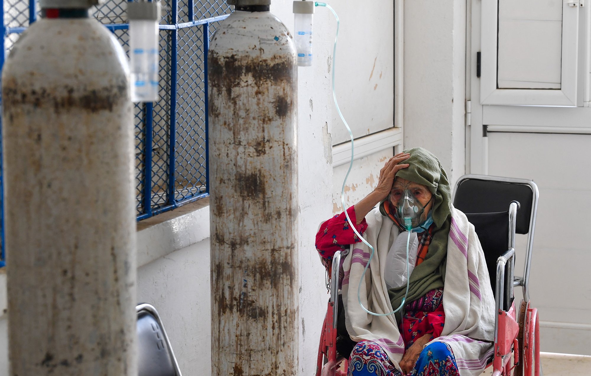 L'image montre une femme âgée assise dans un couloir d'hôpital. Elle porte un masque et une écharpe sur la tête. On peut voir des bouteilles d'oxygène à proximité, indiquant qu'elle reçoit des soins médicaux. L'environnement est simple, avec des murs blancs et des fenêtres qui laissent passer la lumière. L'expression de la femme suggère une certaine inquiétude ou fatigue.
