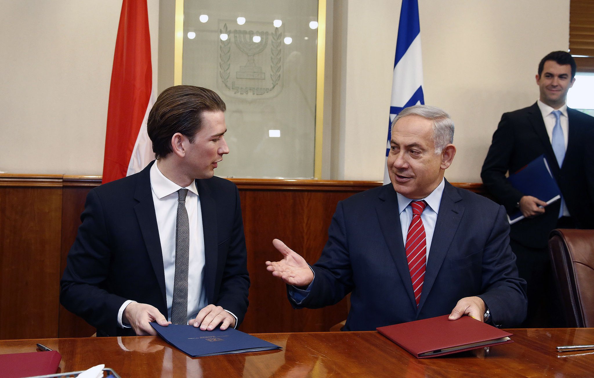 L'image montre deux hommes assis à une table, probablement lors d'une réunion ou d'une cérémonie de signature. L'un d'eux est en costume sombre avec une cravate claire, tandis que l'autre porte une cravate rouge. Ils semblent engagés dans une conversation amicale. À l'arrière-plan, on peut voir des drapeaux, probablement ceux de leurs pays respectifs. L'ambiance générale semble formelle et diplomatique.