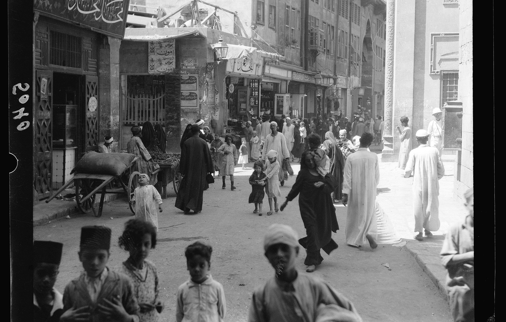 L'image montre une scène animée d'une rue dans une ville. On y voit des groupes de personnes marchant, certaines en conversation. Des enfants jouent au milieu des passants. L'architecture autour d'eux suggère un paysage urbain traditionnel, avec des bâtiments ornés et des boutiques. Il y a aussi une charrette et des femmes et hommes vêtus de vêtements traditionnels. L'ambiance générale semble dynamique et vivante, reflétant la culture locale.