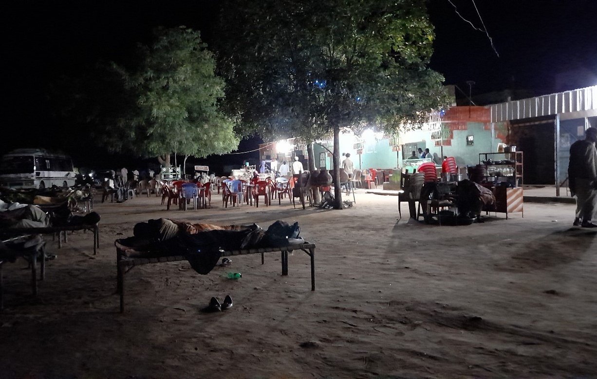 L'image montre un espace en plein air, probablement dans un endroit rural. Il fait nuit, et l'éclairage est principalement fourni par une source lumineuse sur le bâtiment à droite. On peut voir plusieurs tables et chaises disposées, avec des personnes assises et d'autres qui se déplacent. À gauche, il y a des lits ou des matelas disposés sur le sol, suggérant que des gens y dorment ou se reposent. Des arbres sont visibles, ajoutant une touche de verdure à la scène. L'atmosphère semble calme, mais animée par la présence de plusieurs personnes.