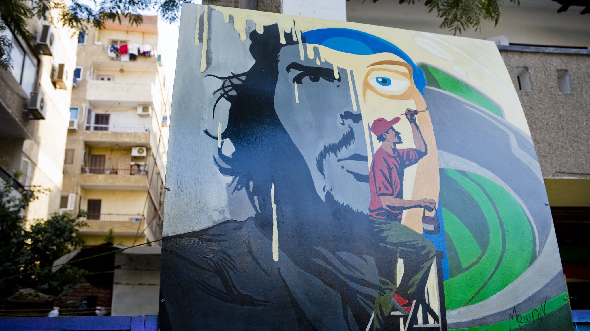 L'image montre une fresque murale colorée. À gauche, il y a le visage d'un homme avec une expression intense, tandis qu'à droite, un artiste est représenté en train de peindre. L'artiste est vêtu d'un pantalon rouge et d'un t-shirt, et il est sur une échelle, concentré sur son œuvre. En arrière-plan, on aperçoit des bâtiments avec des balcons et du linge suspendu. Les couleurs de la fresque sont vives, avec des nuances de bleu et de vert qui contrastent avec les éléments plus sombres du visage.