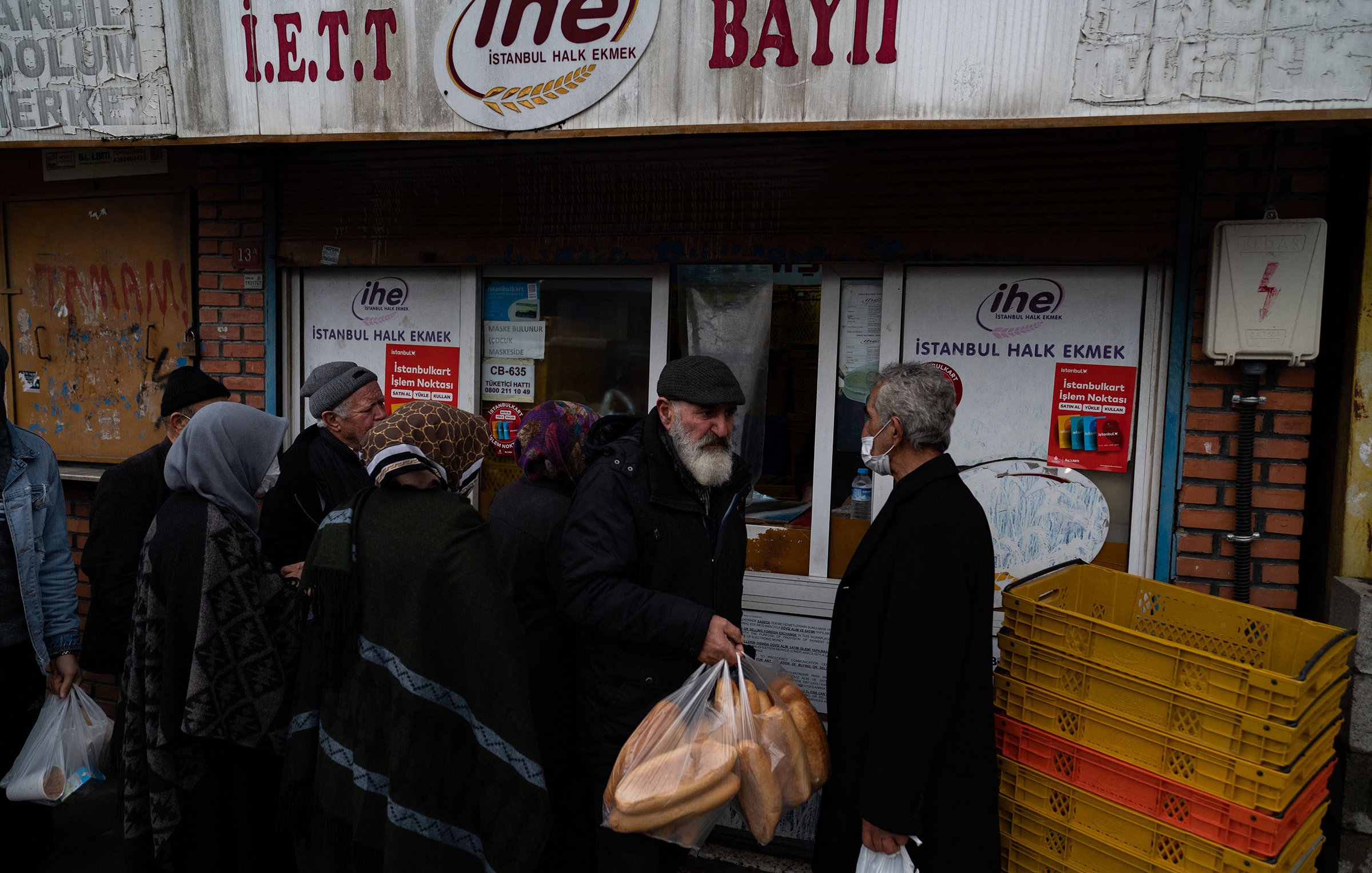 L'image montre une scène animée devant une boulangerie à Istanbul. On aperçoit une file de personnes, majoritairement des personnes âgées, qui attendent pour acheter du pain. Certains portent des sacs en plastique contenant des baguettes. Les gens semblent discuter entre eux, et la boulangerie affiche des panneaux avec des indications sur les produits. L'atmosphère est conviviale et caractéristique des marchés de quartier dans la ville.
