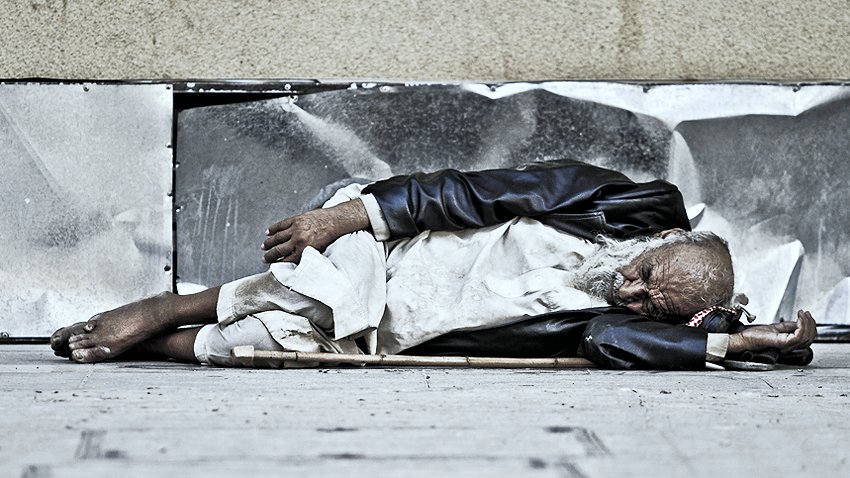 L'image montre un homme âgé allongé sur le sol, semblant endormi. Il porte des vêtements usés et semble être dans une position de repos sur un trottoir. Son visage est marqué par le temps et il est entouré d'un environnement urbain. L'atmosphère évoque une certaine mélancolie et une réflexion sur la vie des personnes sans abri.