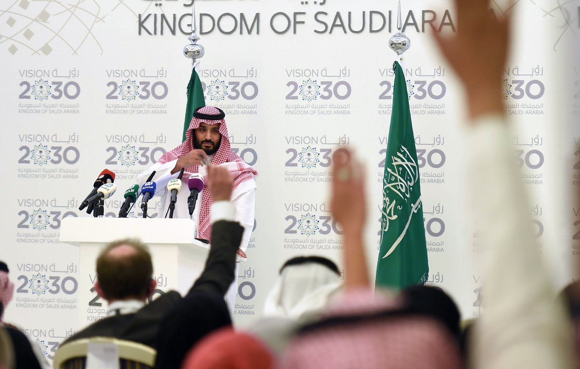 L'image montre une conférence de presse organisée en Arabie Saoudite. Un homme, vêtu d'une robe traditionnelle saoudienne et d'un headdress, se tient devant un podium avec plusieurs microphones. En arrière-plan, on peut voir des drapeaux saoudiens ainsi qu'un panneau affichant des informations sur la Vision 2030 de l'Arabie Saoudite. Plusieurs personnes sont présentes, levant les mains, probablement pour poser des questions. L'atmosphère semble sérieuse, centrée sur des discussions importantes liées à l'avenir du pays.
