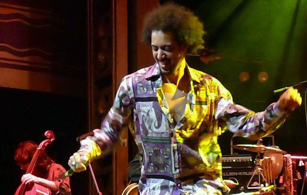 L'image montre un artiste en pleine performance sur scène. Il porte une chemise colorée à motifs et est en train de danser, manifestant une grande énergie. En arrière-plan, on peut apercevoir des musiciens jouant d'instruments, ce qui suggère un concert en direct. L'éclairage de la scène crée une atmosphère dynamique et festive.