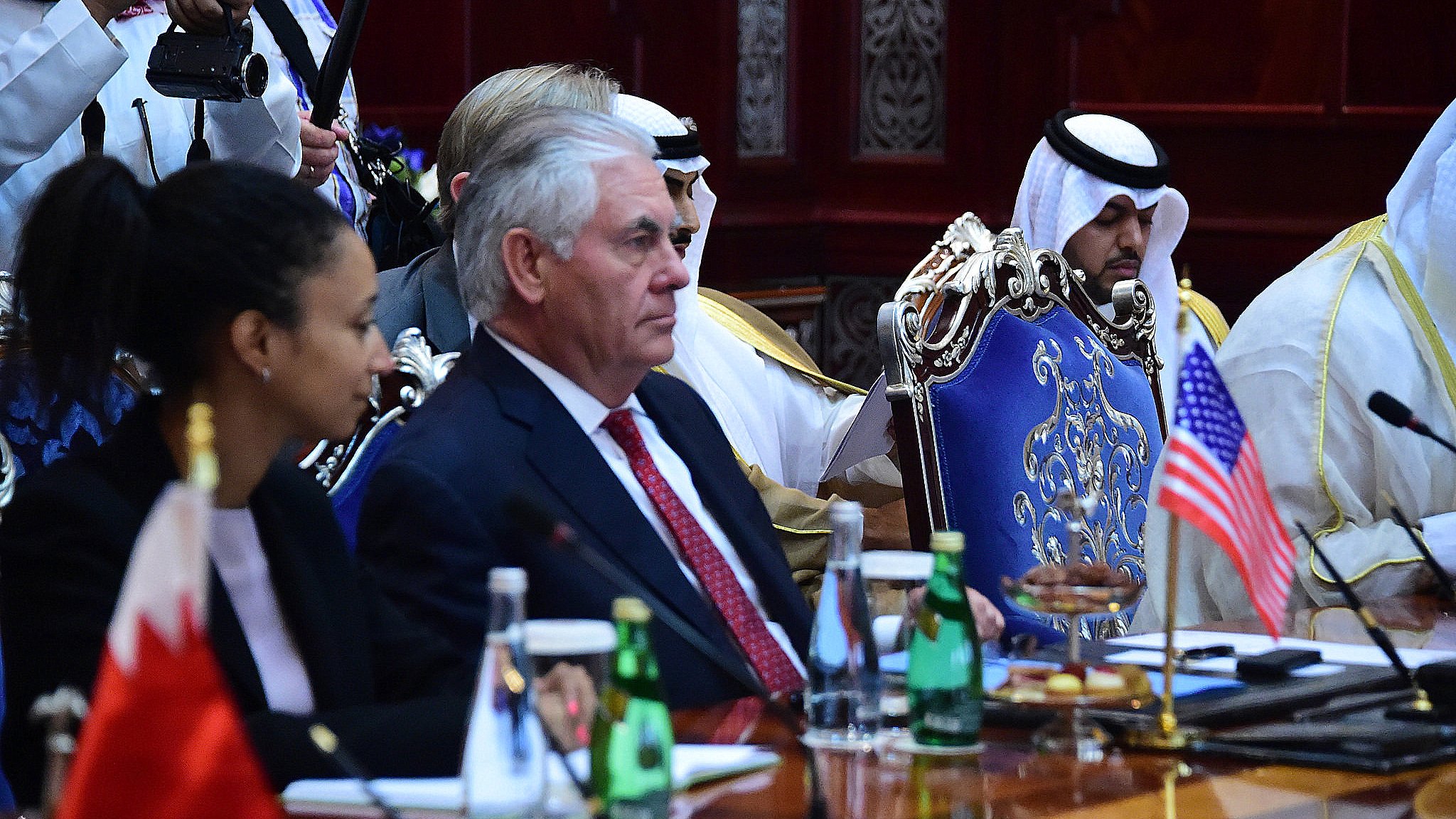 L'image présente une réunion officielle, où l'on peut voir plusieurs personnes assises autour d'une grande table. À gauche, une femme est accoudée à la table, tandis qu'à droite, un homme en costume, probablement un diplomate, semble écouter attentivement. Au centre, il y a un drapeau américain, et derrière, d'autres participants portant des vêtements traditionnels peuvent être aperçus. L'environnement semble formel, avec une décoration riche et des bouteilles d'eau sur la table.