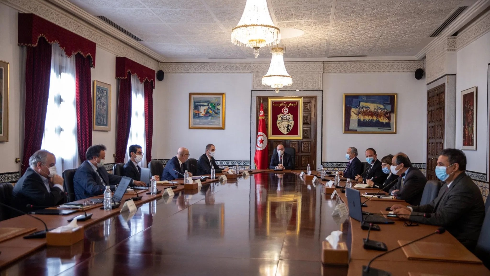 L'image montre une réunion qui se déroule dans une salle de conférence élégante. Il y a plusieurs personnes assises autour d'une grande table en bois. Au centre, un homme semble diriger la réunion, tandis que les autres participants l'écoutent attentivement. La salle est décorée de tableaux et présente des fenêtres avec des rideaux rouges. Tous les participants portent des masques, ce qui suggère une prudence sanitaire. L'atmosphère est formelle, typique d'une réunion gouvernementale ou d'affaires.