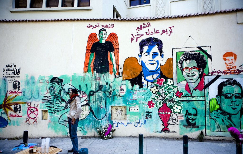 L'image montre un mur recouvert de fresques murales colorées. On y voit plusieurs portraits et illustrations. L'une des figures centrales semble représenter une personne avec des ailes, symbole souvent associé à un message d'espoir ou de mémoire. Des textes en arabe et en français peuvent être aperçus, évoquant des thèmes de la liberté et de la résistance. En bas, une personne se tient debout, probablement en train de peindre ou d'admirer l'œuvre. L'atmosphère générale dégage une impression vibrante et engagée, reflétant une culture urbaine riche.