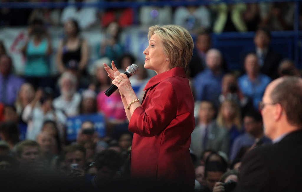 L'image montre une femme prenant la parole devant un public. Elle porte un manteau rouge et utilise un microphone pour s'exprimer. En arrière-plan, on peut voir une foule de personnes, certaines tenant des panneaux ou des affiches. L'éclairage et l'ambiance suggèrent un événement politique ou une réunion publique.