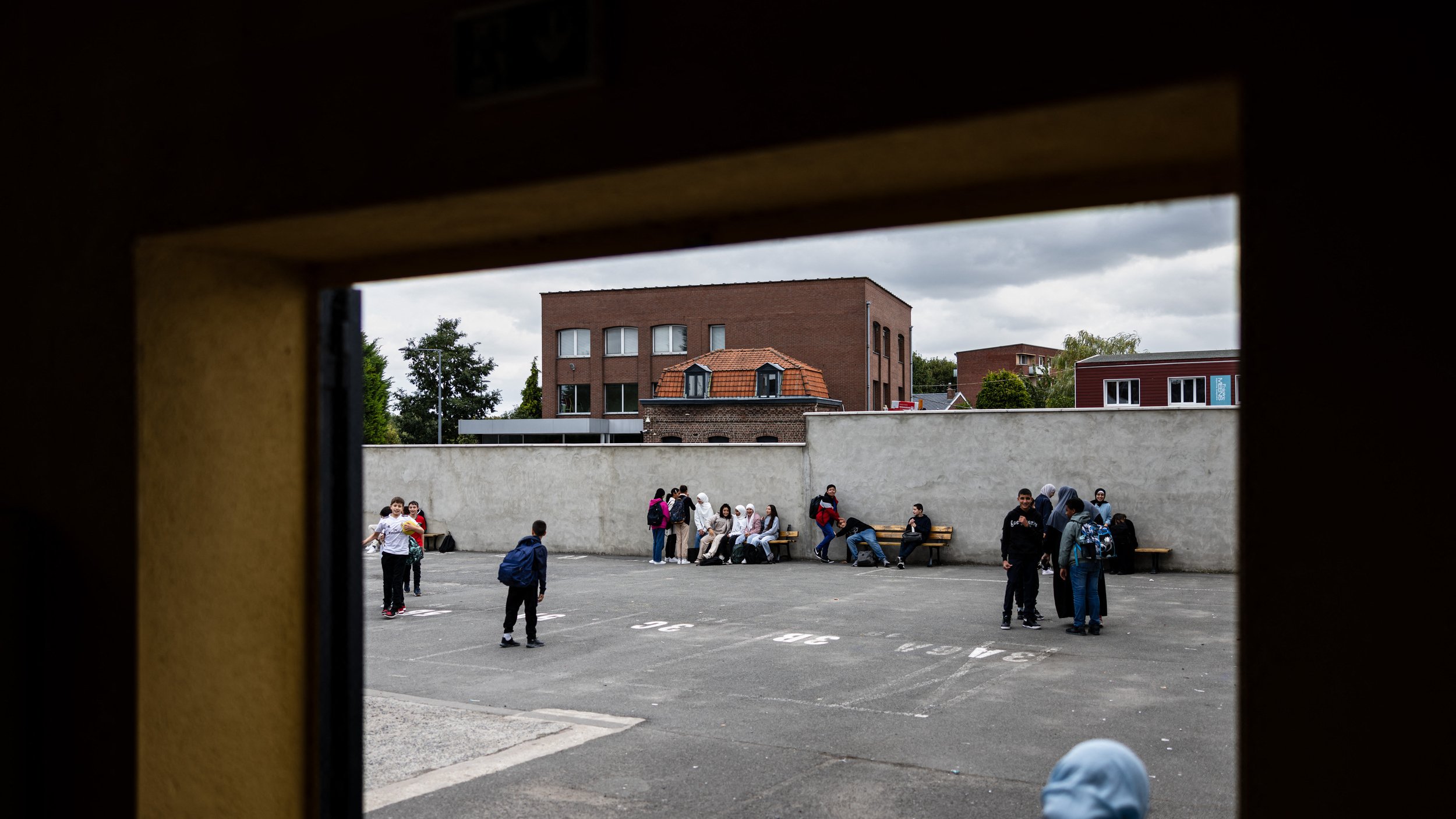 L'image montre une scène d'un espace extérieur, probablement une cour de récréation ou un terrain de sport. On peut voir plusieurs groupes de jeunes, répartis sur le sol, assis ou discutant ensemble. À l'arrière-plan, il y a des bâtiments avec des fenêtres et un toit de briques. Le ciel est nuageux, ce qui donne une ambiance un peu sombre à la scène. Les jeunes semblent engagés dans des conversations ou des activités sociales.