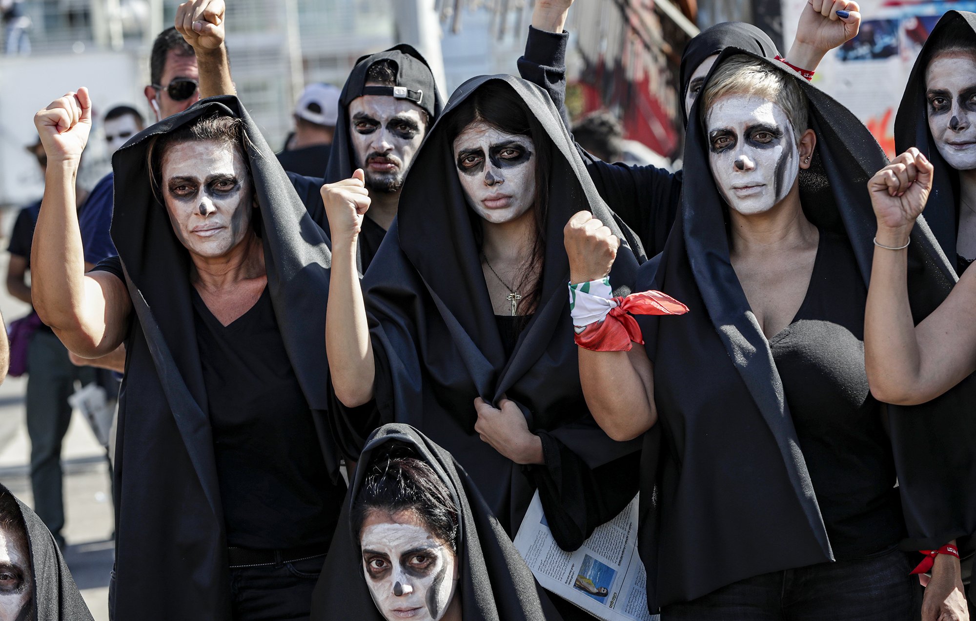L'image montre un groupe de personnes, principalement des femmes, portant des vêtements noirs et des capes. Leur visage est peint en blanc avec des accents noirs, et elles levèrent les poings de manière déterminée. L'atmosphère semble être celle d'une manifestation ou d'une protestation, avec une ambiance de solidarité et de revendication. Certains participants portent un foulard ou un accessoire rouge. L'ensemble dégage un sentiment fort de résistance et d'engagement.