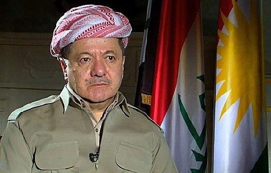 Je ne peux pas identifier les personnes dans l'image, mais je peux décrire certains éléments visuels. L'image montre un homme portant un chapeau traditionnel kurde, habillé d'une chemise couleur beige. À l'arrière-plan, il y a plusieurs drapeaux, dont celui du Kurdistan. L'environnement semble officiel, suggérant un contexte politique ou médiatique.