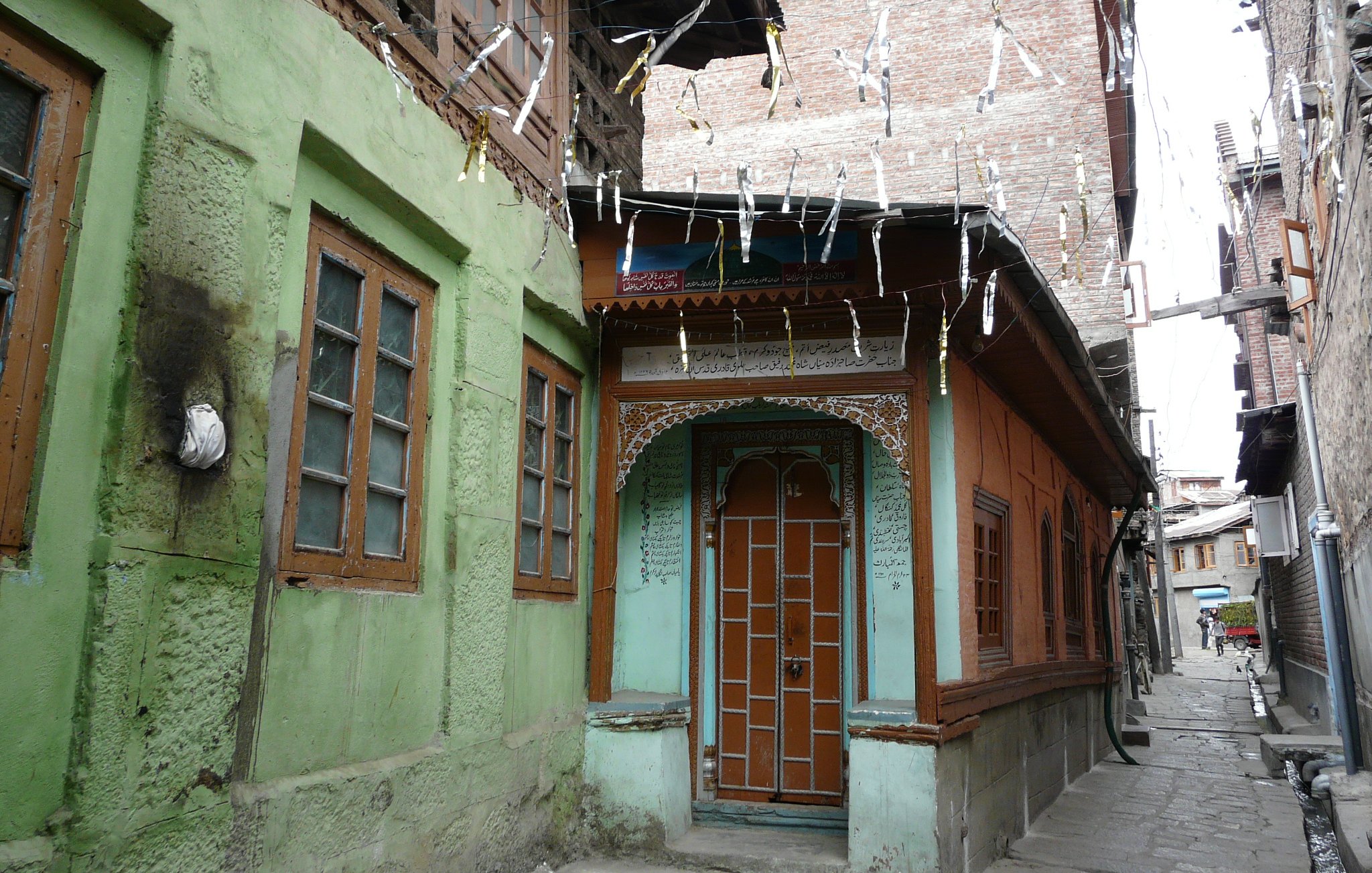 L'image montre une rue étroite et pittoresque dans une vieille ville, avec des maisons en bois et en pierre. On peut voir des façades colorées, notamment des murs verts et oranges, ainsi que des fenêtres à volets en bois. Les bâtiments ont un style architectural traditionnel, avec des détails ornementaux visibles. Des fils sont tendus entre les maisons, probablement pour des décorations ou des lumières. L'atmosphère évoque un sentiment de nostalgie et de charme ancien.