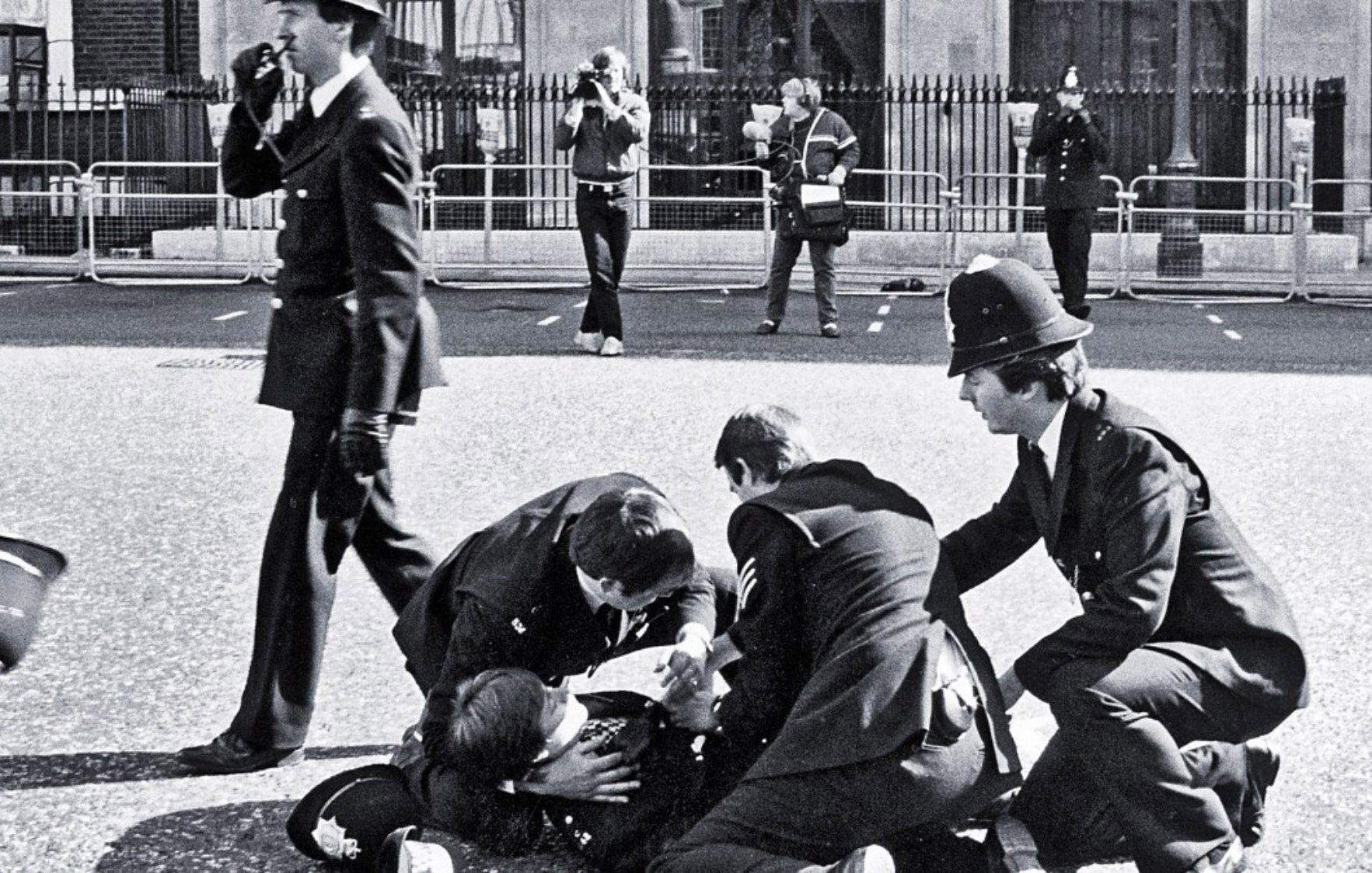 L'image montre une scène policière en noir et blanc. Plusieurs policiers en uniforme entourent une personne au sol, visiblement en train d'être arrêté ou assisté. D'autres policiers se tiennent en arrière-plan, certains prenant des notes ou observant la situation. L'environnement semble urbain, avec des barrières de sécurité visibles, probablement autour d'une zone publique. L'atmosphère dégage une impression d'urgence et d'autorité.