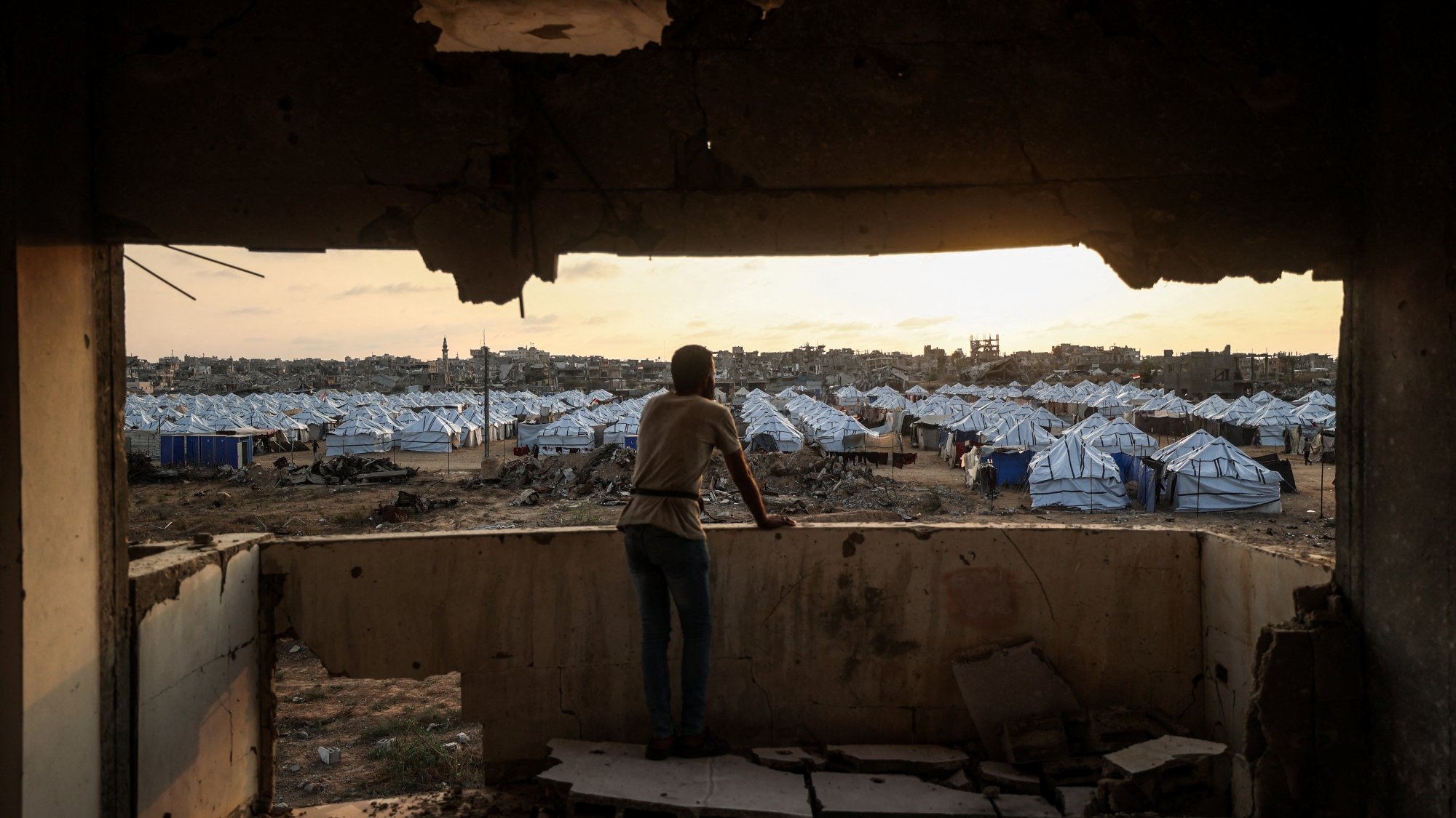 Un jeune observe un camp de tentes depuis une fenêtre endommagée au crépuscule.