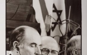 L'image montre deux hommes assis à une table lors d'une conférence. L'un des hommes, portant des lunettes et un costume, semble être en train de parler, tandis que l'autre, avec un regard sérieux, écoute attentivement. En arrière-plan, des drapeaux sont visibles, y compris un drapeau israélien. L'atmosphère semble formelle, ce qui suggère que l'événement a une importance politique ou diplomatique.