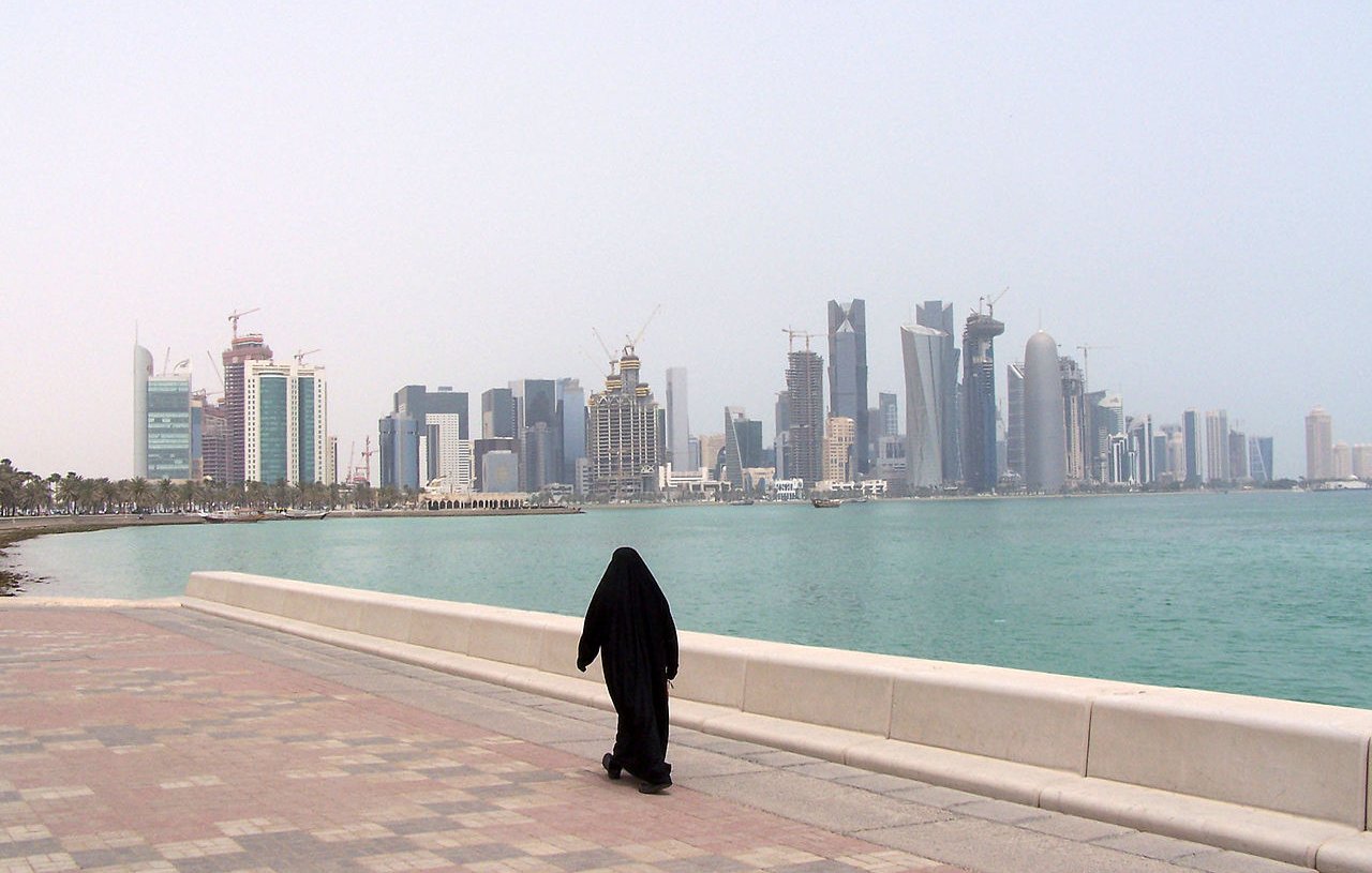 L'image montre une personne vêtue d'une tenue traditionnelle noire, marchant le long d'une promenade au bord de l'eau. En arrière-plan, on aperçoit une ligne d'horizon urbaine avec des gratte-ciel modernes et des bâtiments en construction, représentant une ville côtière. L'ambiance est calme, avec un ciel légèrement brumeux et une mer tranquille. Les pavés de la promenade ajoutent une touche de couleur à la scène.