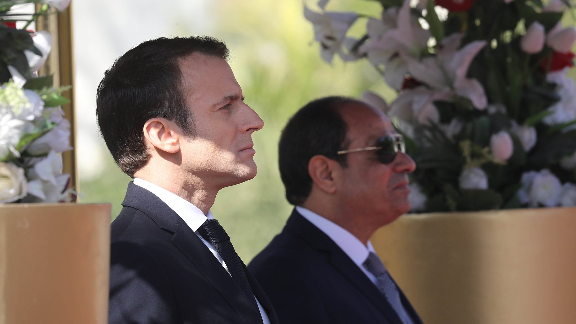 L'image montre deux hommes debout, apparemment lors d'un événement officiel. L'un d'eux est en costume sombre avec une cravate, il a une expression concentrée et regarde droit devant lui. L'autre homme, également en costume, porte des lunettes de soleil et semble observateur. Ils se tiennent près de décorations florales, suggérant un cadre cérémonieux.