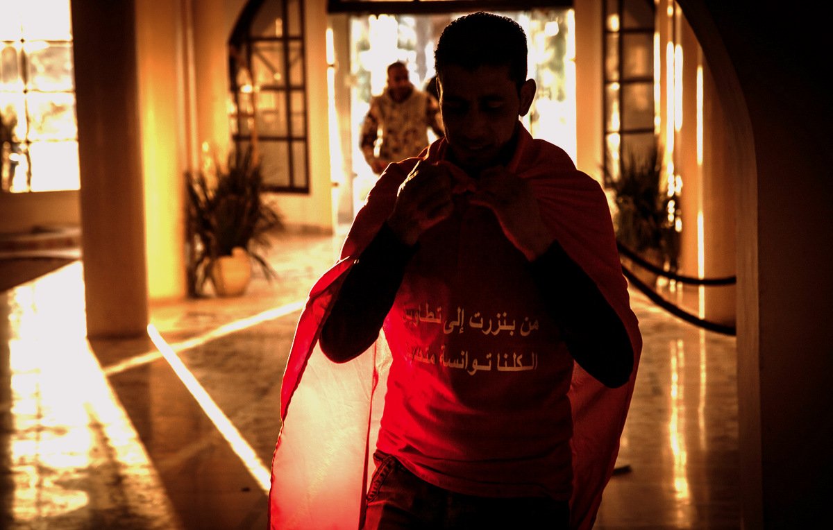 L'image montre un homme portant un t-shirt rouge avec un drapeau. Il semble s'apprêter à sortir, avec une ambiance chaleureuse et lumineuse en arrière-plan. On peut voir des éléments de décor qui suggèrent un espace intérieur, avec des silhouettes floues et une lumière dorée qui crée une atmosphère dramatique. L'homme porte une expression sérieuse et déterminée.