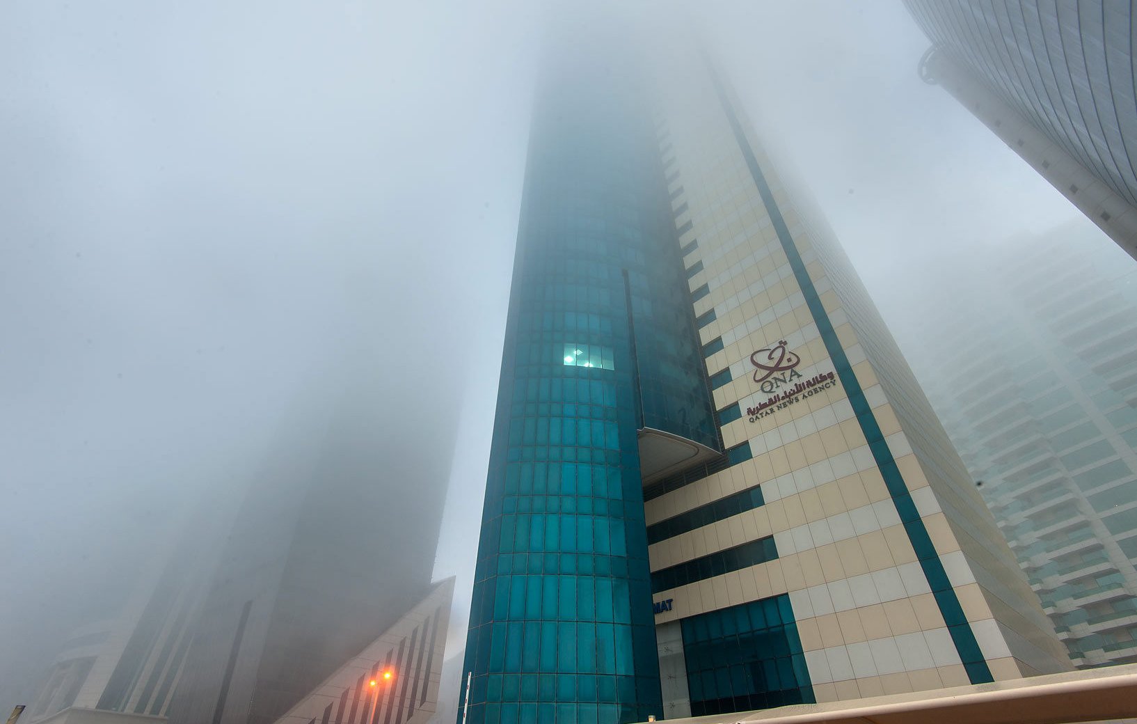 L'image montre un paysage urbain avec des gratte-ciels partiellement cachés par un épais brouillard. La visibilité est réduite, ce qui donne une atmosphère mystérieuse et peu lumineuse. Les bâtiments ont des façades modernes, avec des reflets de verre. On peut distinguer des éléments architecturaux, mais l'intensité du brouillard rend difficile l'observation claire des détails. Les lumières des rues sont visibles, créant un contraste avec l'environnement brumeux.