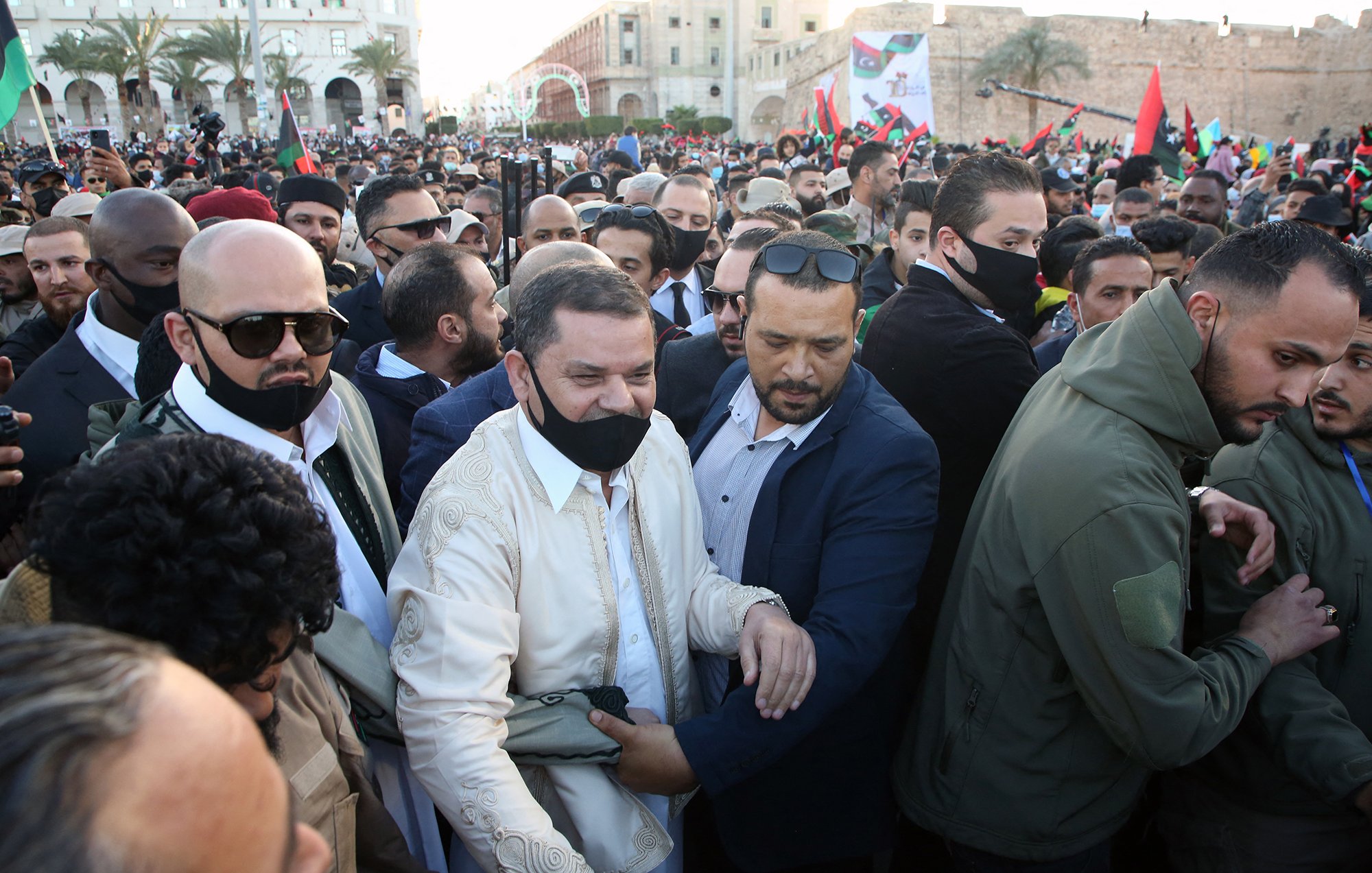 L'image montre une foule dense, avec un groupe de personnes rassemblées lors d'un événement. Au centre, un homme en tenue traditionnelle est entouré de plusieurs personnes portant des masques. L'ambiance semble être celle d'un rassemblement ou d'une célébration. Les participants affichent des expressions d'enthousiasme, certains tenant des bannières ou des drapeaux. En arrière-plan, des bâtiments et de la verdure sont visibles, ce qui indique une atmosphère festive.