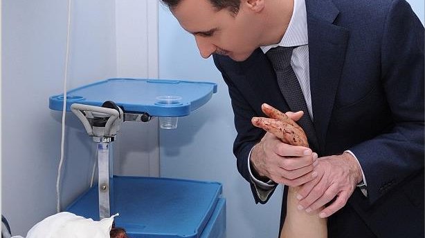 L'image montre un homme en costume qui se penche vers une personne allongée sur un lit d'hôpital, avec un bandage sur la tête. La personne blessée a une expression de souffrance, et l'homme semble lui parler ou lui apporter du soutien, tenant son bras. L'environnement est médical, avec des équipements visibles en arrière-plan.