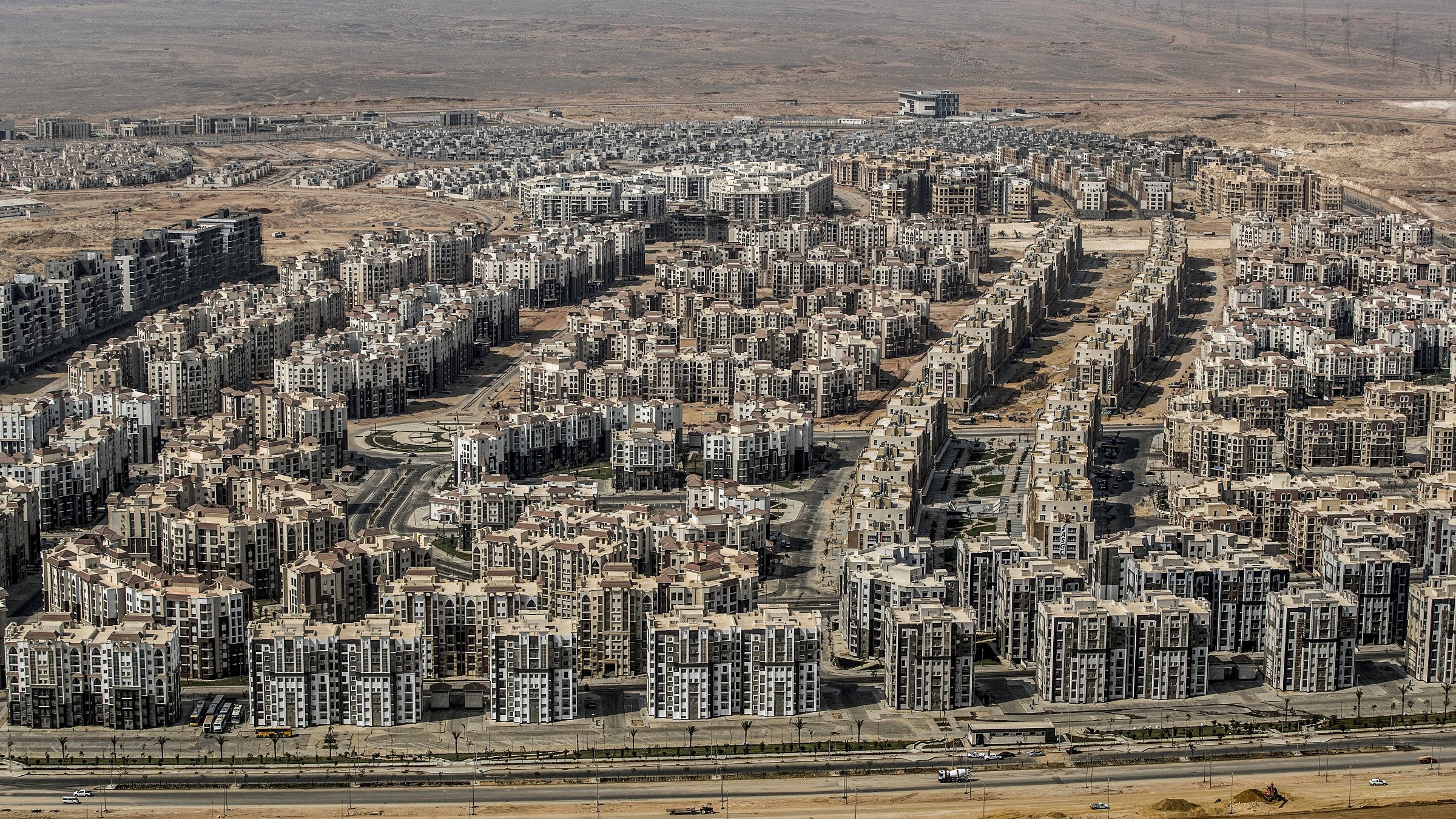 L'image montre une vaste zone urbaine composée de nombreux immeubles de plusieurs étages, organisés de manière dense. On peut voir des routes qui traversent ces secteurs résidentiels, avec des infrastructures modernes. En arrière-plan, le paysage est désertique, ce qui évoque un contraste entre l'urbanisation et l'environnement naturel environnant. L'ensemble dégage une impression de développement urbain important dans une région aride.