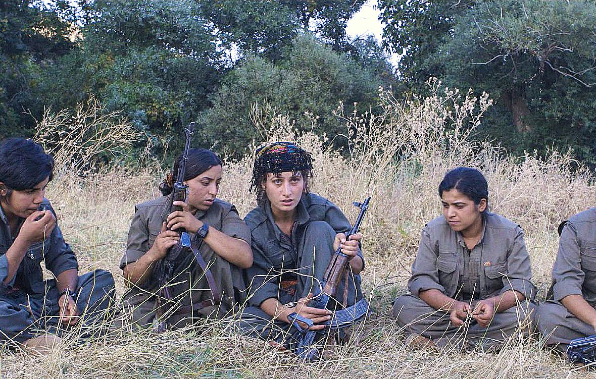 L'image montre un groupe de cinq femmes assises dans une prairie, entourées de végétation. Elles portent des vêtements militaires et semblent concentrées sur une activité commune. Certaines d'entre elles tiennent des armes à feu, tandis que d'autres ont les mains posées sur leurs genoux. Leurs expressions faciales et leur posture suggèrent une ambiance sérieuse et déterminée.