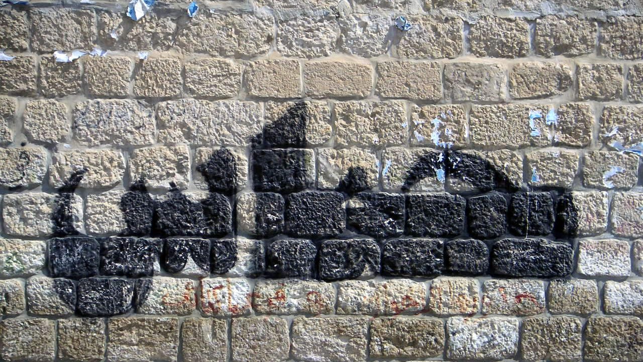 L'image montre un mur de pierres avec un graffiti en forme de silhouette noire. Le mur semble ancien et légèrement abîmé, avec des morceaux de papier déchirés. La silhouette créée par le graffiti évoque des formes humaines, mais sans détails précis. Les pierres du mur sont bien visibles et ajoutent une texture rustique à l'ensemble.