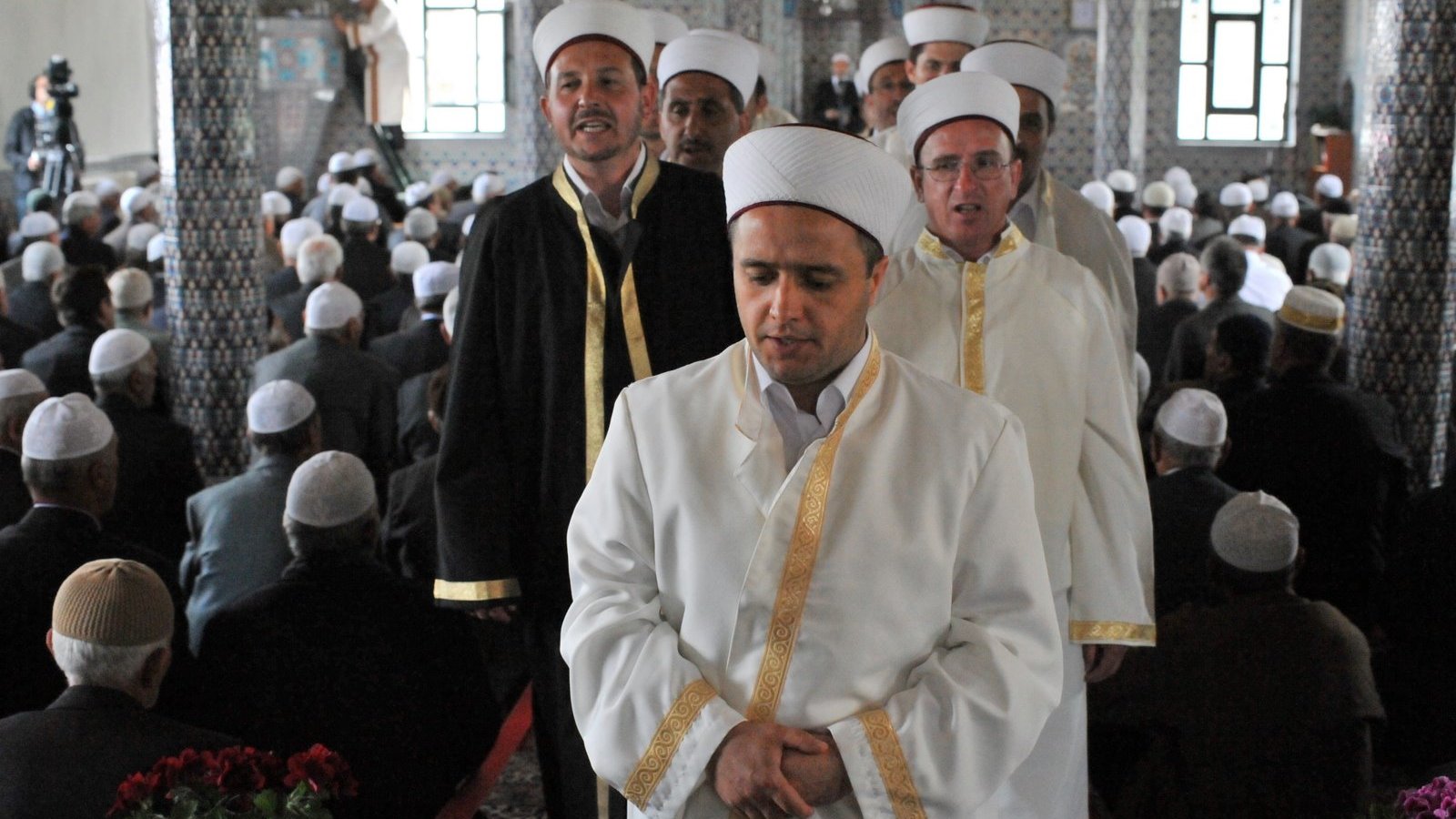 L'image montre un groupe d'hommes portant des vêtements traditionnels, notamment des robes blanches et des turbans. Ils semblent être en procession, probablement lors d'un événement religieux. En arrière-plan, de nombreuses personnes sont assises, probablement en prière ou en attente d'une cérémonie. L'architecture de la pièce inclut des motifs décoratifs et des fenêtres qui laissent entrer la lumière, créant une atmosphère solennelle.