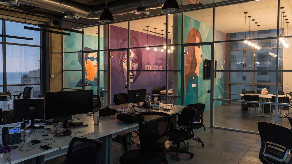 L'image montre un espace de bureau moderne avec de grandes fenêtres offrant une vue sur l'extérieur. Les murs sont décorés de peintures murales colorées représentant des visages stylisés. Le mobilier est minimaliste et fonctionnel, avec des tables de travail et des chaises ergonomiques. L'éclairage est tamisé, créant une ambiance chaleureuse malgré le design contemporain. On peut également voir des ordinateurs sur les bureaux, indiquant que l'espace est utilisé pour le travail.