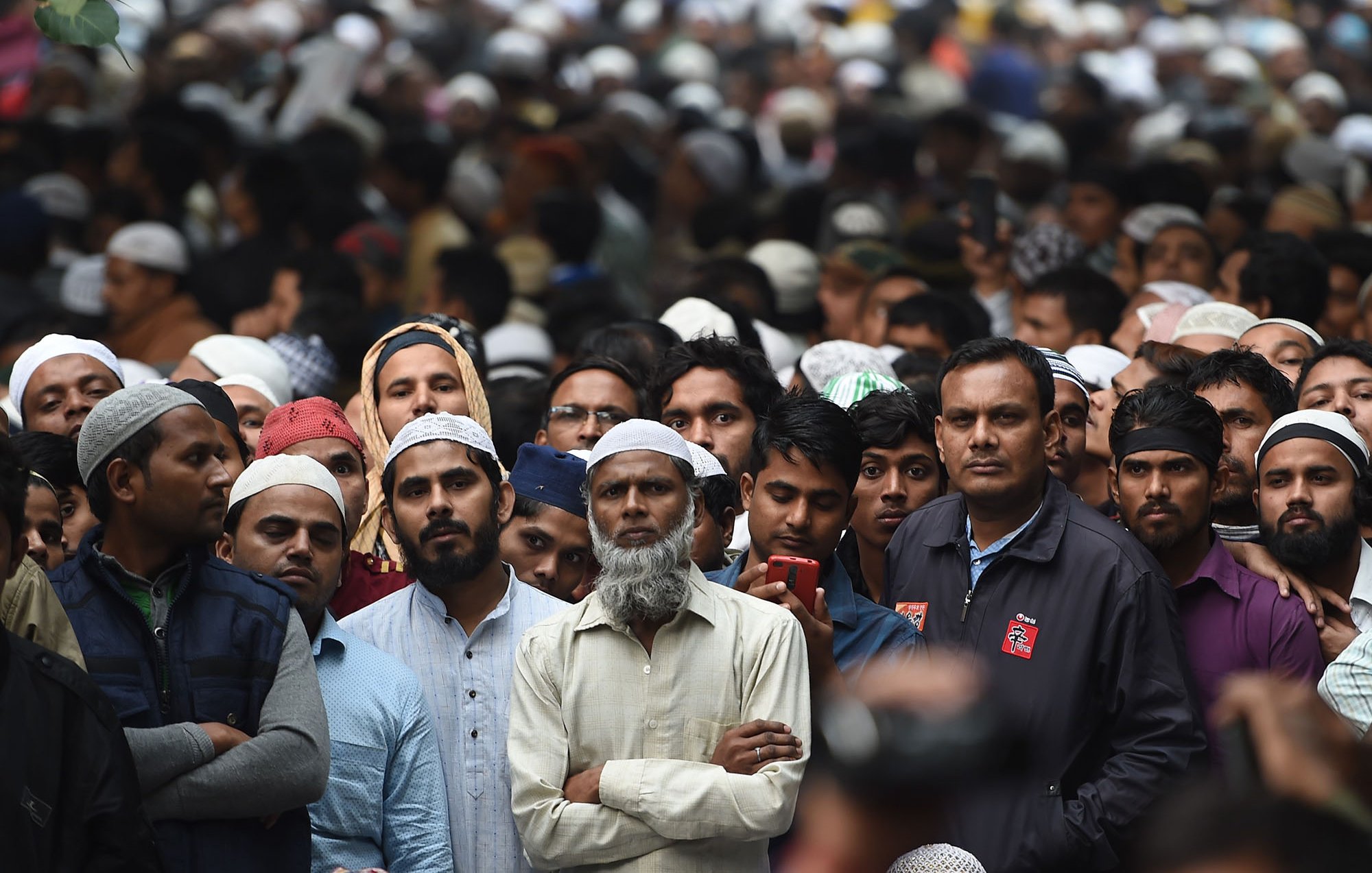 L'image montre une grande foule d'hommes, principalement vêtus de vêtements traditionnels, avec plusieurs d'entre eux portant des couvre-chefs. Les expressions des gens varient, certains semblent attentifs, tandis que d'autres regardent vers l'avant. L'ambiance dégage un sentiment de concentration et de rassemblement, possiblement pour un événement ou une cérémonie.