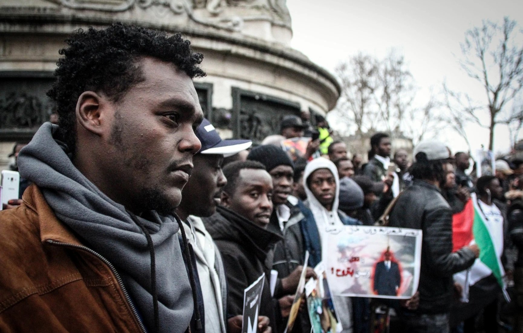 L'image montre un groupe de personnes rassemblées, probablement lors d'une manifestation ou d'un événement public. Au premier plan, un homme au port de tête pensif se distingue, entouré d'autres participants qui tiennent des pancartes et des photos. L'atmosphère semble engagée et solidaire, avec des affiches qui pourraient exprimer des revendications ou des messages de protestation. L'environnement urbain est visible en arrière-plan, suggérant que cet événement se déroule dans un espace public.