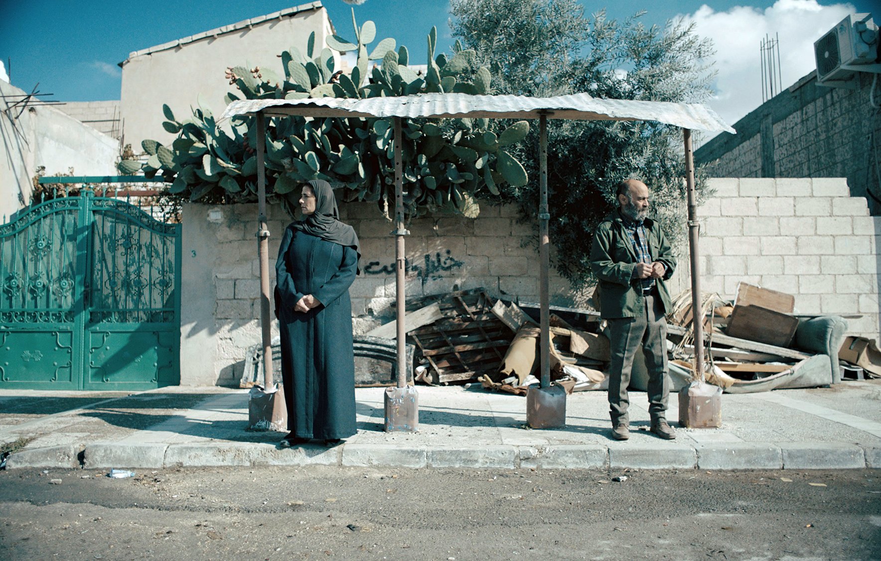 L'image montre une scène dans une rue, où deux personnes se tiennent sous un abri. À gauche, une femme est vêtue d'une longue robe sombre et porte un foulard, tandis qu'à droite, un homme, habillé de manière militaire, se tient debout, les mains derrière le dos. En arrière-plan, on voit un mur en briques avec des débris et des plantes, notamment un cactus. L'environnement a une ambiance un peu désolée et montre des signes d'abandon. Le ciel est partiellement nuageux, ajoutant une certaine atmosphère à la scène.