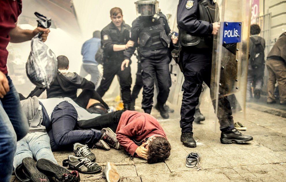 L'image montre une scène de manifestation ou d'émeute. On peut voir plusieurs personnes au sol, certaines semblant blessées ou cernées par des forces de police. Des policiers en tenue anti-émeute avancent dans une atmosphère de chaos, avec des gaz lacrymogènes obscurcissant l'air. L'environnement paraît tendu, témoignant d'un conflit entre manifestants et autorités.