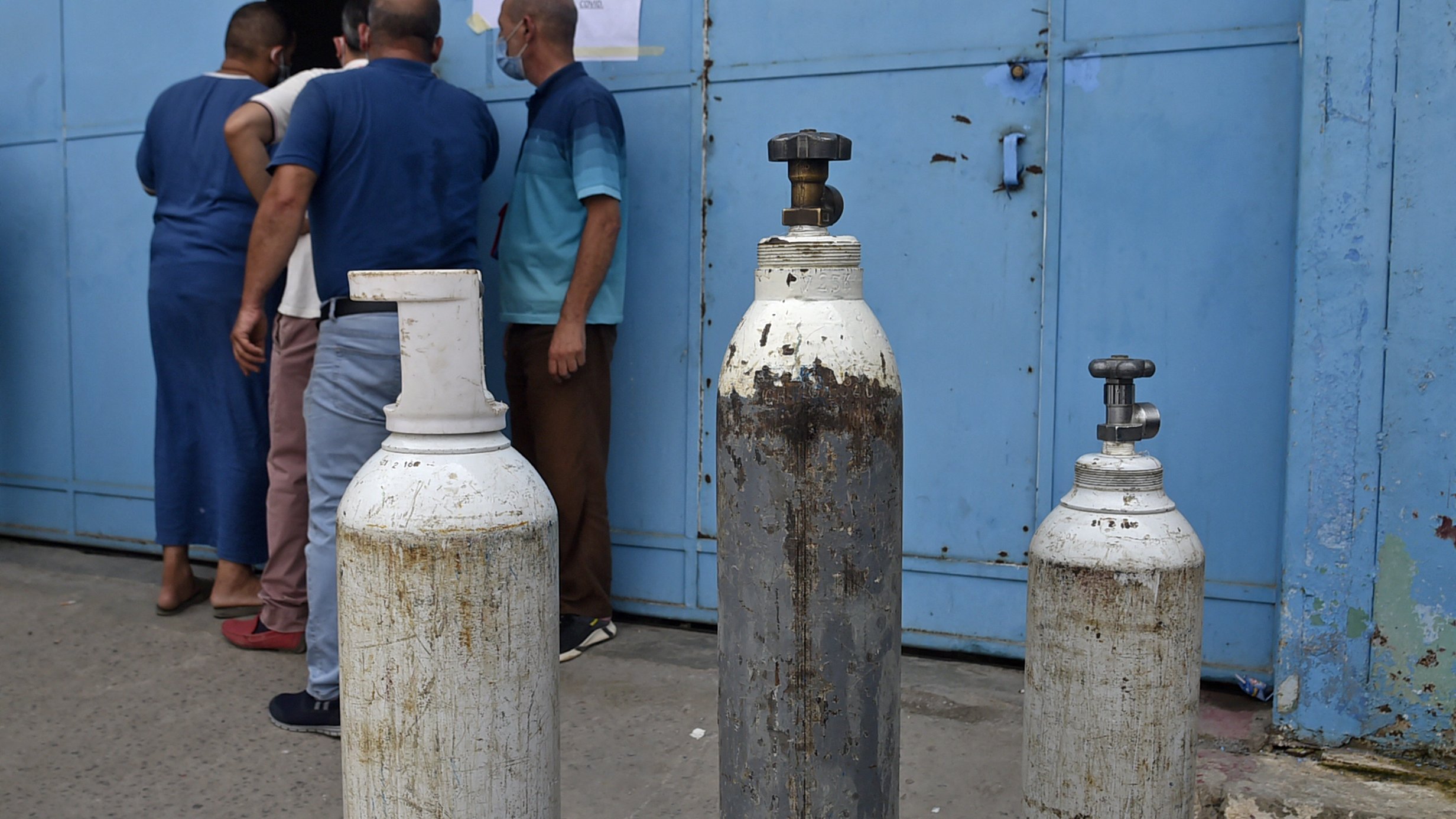 L'image montre un groupe de personnes devant une porte bleue. Sur le devant, il y a plusieurs bouteilles de gaz, dont certaines sont blanches et d'autres en métal. Les bouteilles semblent usées et peuvent être utilisées pour stocker des gaz, comme l'oxygène. Les personnes à l'arrière semblent attendre ou discuter, tandis que l'environnement semble être un lieu urbain.