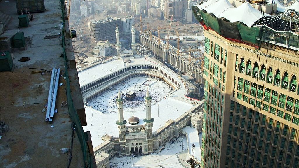 L'image montre une vue aérienne de la Kaaba à La Mecque, en Arabie Saoudite. On peut voir le complexe du Haram qui comprend la mosquée entourant la Kaaba. Il y a de nombreux pèlerins qui s'y trouvent, et l'environnement urbain avec des bâtiments modernes en arrière-plan. On aperçoit également des travaux de construction en cours sur le côté, suggérant une expansion des infrastructures autour du site sacré. Le ciel est légèrement brumeux, ce qui donne une ambiance particulière à la scène.