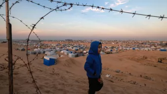 L'image montre un jeune garçon portant une veste bleue qui marche dans un paysage désertique. En arrière-plan, on aperçoit un camp de réfugiés avec de nombreuses tentes et abris disposés sur le sol sablonneux. Un barbelé est visible au premier plan, ajoutant une dimension de sécurité ou de séparation à la scène. Le ciel est partiellement dégagé, avec des teintes de bleu et d'orange, suggérant peut-être la fin de la journée. L'ensemble transmet une atmosphère à la fois de vulnérabilité et d'espoir.