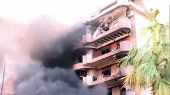 L'image montre un bâtiment en proie à un incendie. De la fumée noire s'échappe des fenêtres, tandis que l'extérieur du bâtiment semble endommagé. Des éléments de végétation, comme un palmier, sont visibles au premier plan, contrastant avec la scène chaotique et la destruction. Le ton général de l'image est sombre, mettant en évidence l'urgence et la gravité de la situation.