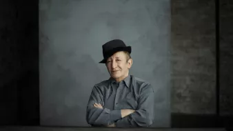 L'image montre un homme assis avec un chapeau noir. Il a un léger sourire et les bras croisés sur une table. Il porte une chemise grise. L'arrière-plan est simple, avec une texture de mur gris qui crée une atmosphère sobre et contemplative. La lumière est douce, accentuant les traits de son visage.
