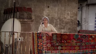 L'image montre une femme se tenant sur un balcon, regardant au loin. Elle porte un haut clair et un foulard sur la tête. Derrière elle, un grand tapis coloré est accrochée à la balustrade, avec des motifs détaillés et vibrants. En arrière-plan, on aperçoit un mur et une antenne satellite, ce qui suggère un environnement urbain. L'éclairage donne une atmosphère chaleureuse à la scène.