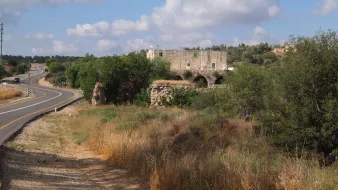 L'image montre un paysage rural avec une route sinueuse sur la gauche. À droite, il y a des herbes hautes et quelques arbres. Au fond, on aperçoit des ruines d'un bâtiment en pierre, probablement une ancienne construction, entourées de verdure. Le ciel est légèrement nuageux, créant une atmosphère paisible et naturelle.
