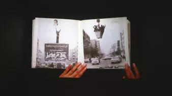 L'image montre un livre ouvert, tenu par des mains, avec des pages affichant des photos en noir et blanc. L'une des pages présente une grande pancarte ou affiche, tandis que l'autre illustre une rue avec des voitures. L'ensemble évoque des thèmes de mouvement, de protestation ou d'événements historiques, en jouant sur le contraste entre les images et le texte visibles. L'arrière-plan est sombre, ce qui met en valeur le livre et les illustrations qu'il contient.