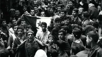 L'image montre une foule dense de personnes rassemblées dans une rue. Au premier plan, plusieurs individus sont visibles, dont un homme qui semble porter un drapeau avec un symbole distinctif. Les expressions des visages varient, certains levant les bras en signe de soutien ou d'enthousiasme. On peut observer des jeunes, des adultes et même des enfants parmi la foule. L'atmosphère semble intense et chargée d'émotion, indiquant un moment significatif ou une manifestation. La scène est en noir et blanc, ce qui lui donne un aspect historique.